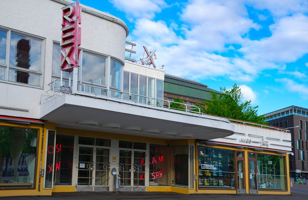 Vintage Rex cinema building with bold red signage and art deco canopy surrounded by green trees.