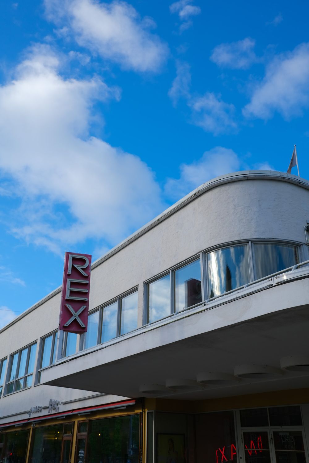 Art Deco Rex cinema building with curved white facade, large windows, and bold red REX sign under a blue sky.