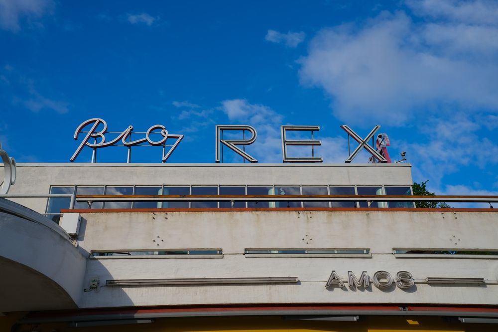 Vintage Bio Rex cinema sign with neon letters against a blue sky, featuring retro architecture and AMCS branding.