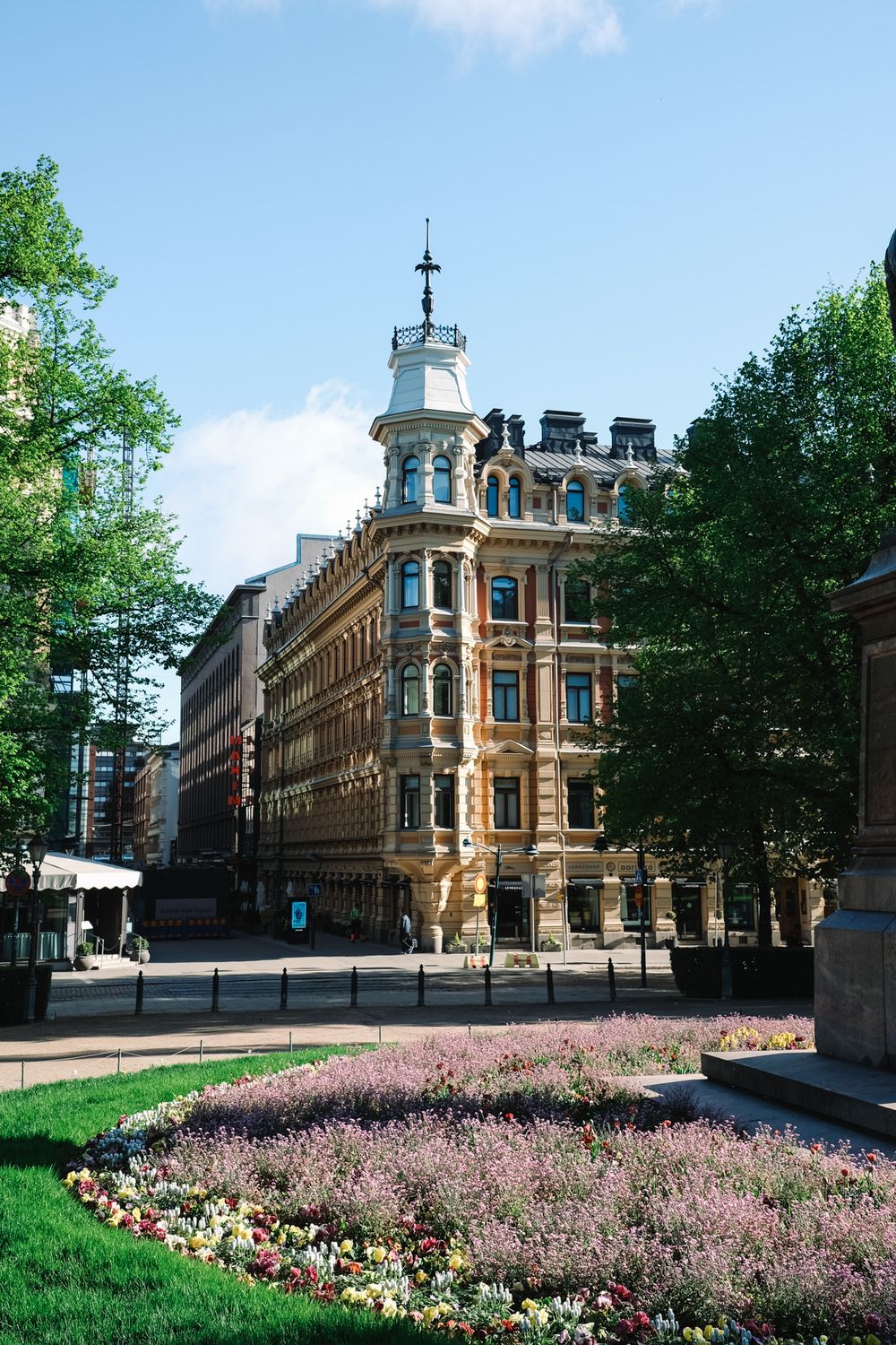 An ornate Victorian corner building with a turret tower stands beside colorful flower beds in a sunny Helsinki square.