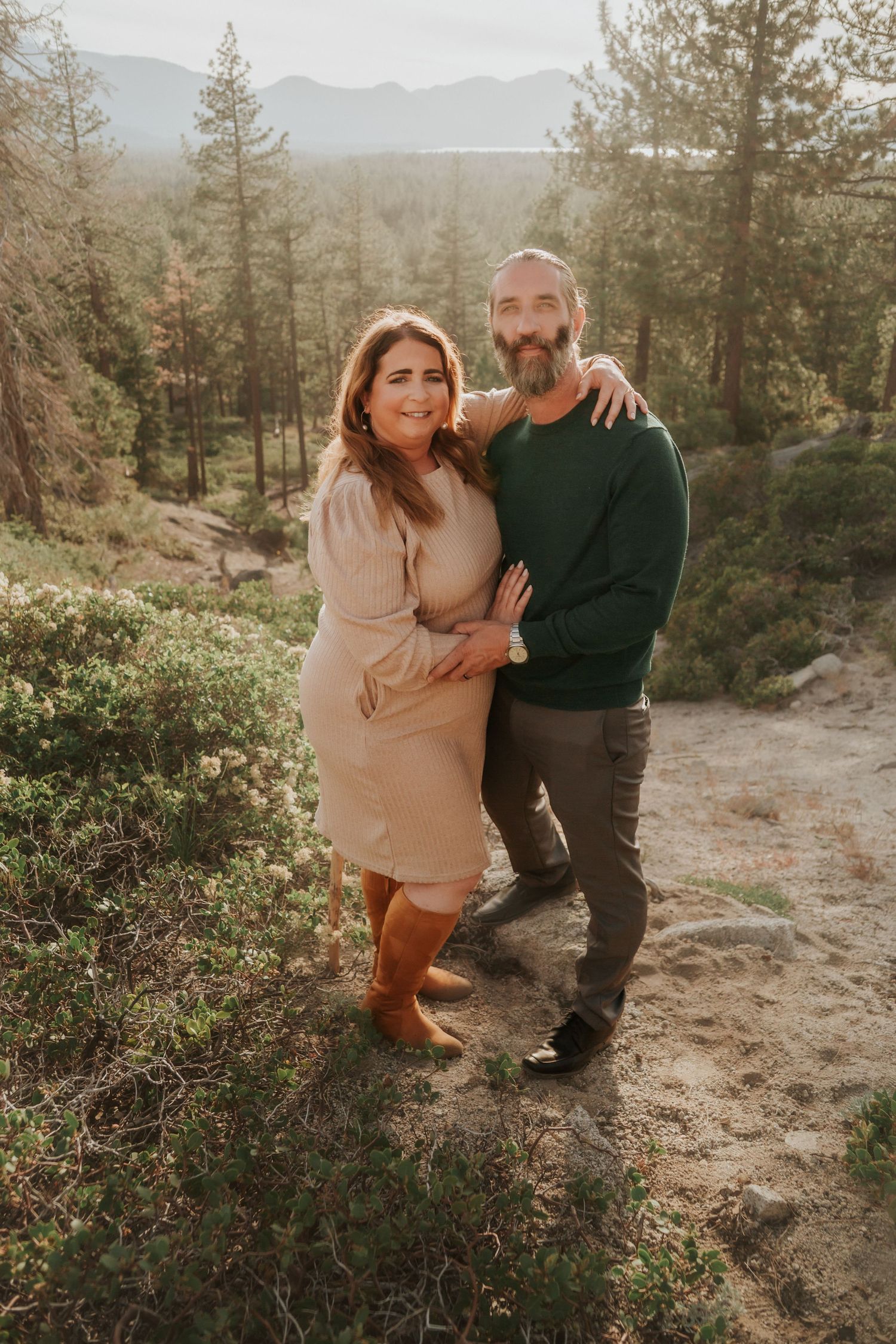 Smiling couple embracing in a sunlit pine forest setting.