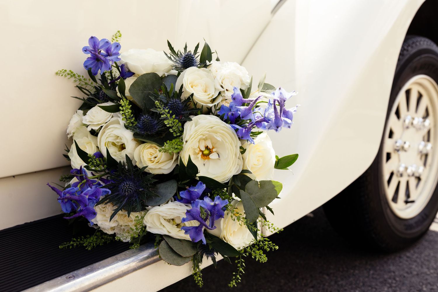 Elegant wedding car bouquet featuring white roses and blue flowers attached to a cream vintage vehicle.