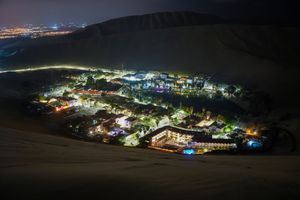 Aerial night view of a glowing desert oasis town nestled between sand dunes with city lights in the distance.