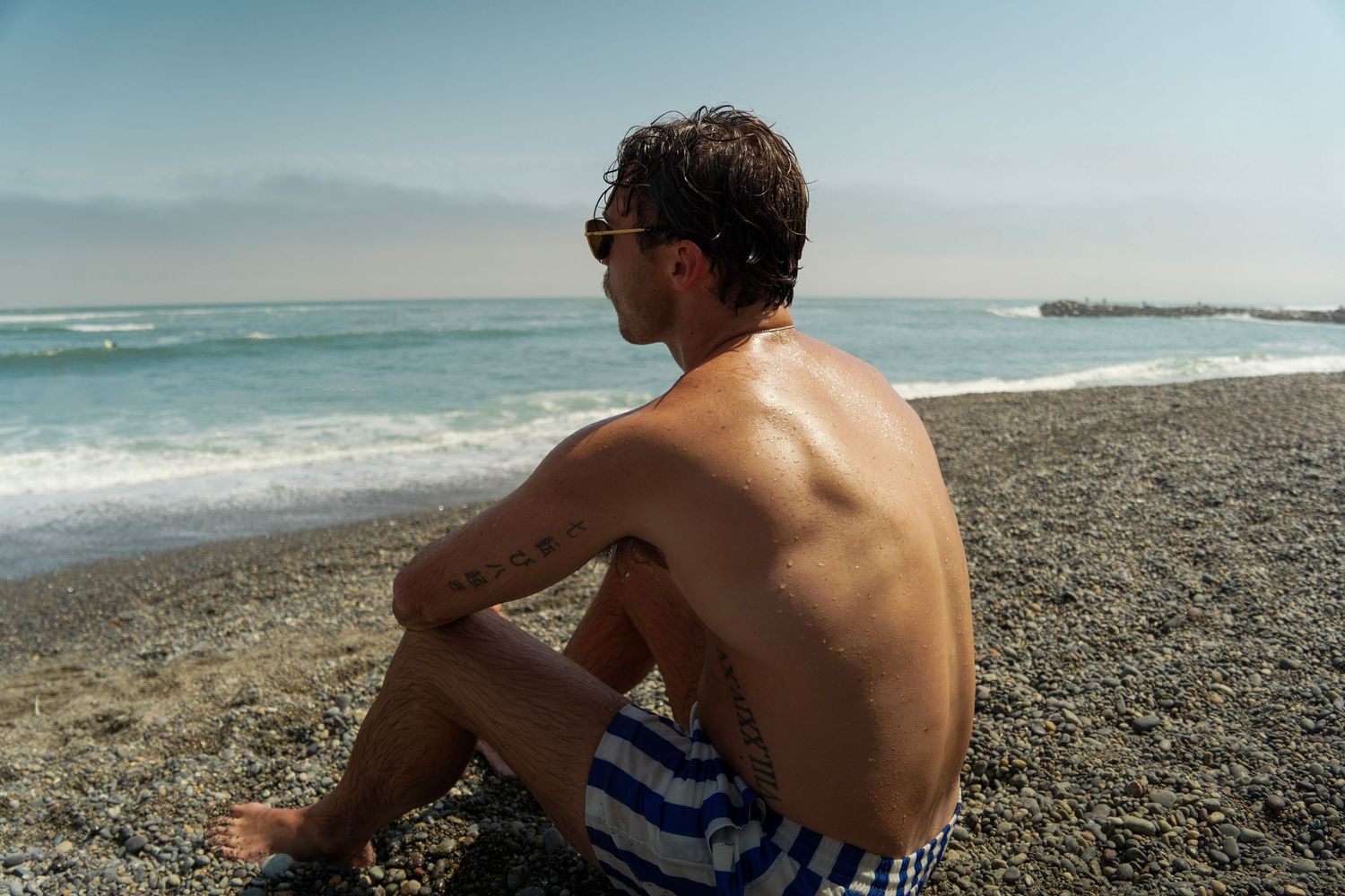 Shirtless man in striped swim trunks sitting on a pebble beach gazing at ocean waves under a clear sky.