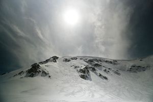 Snow-covered mountain peak under dramatic cloudy sky with sun breaking through, showcasing alpine winter landscape.