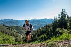 Trail runner with trekking poles ascending a mountain path with scenic green valley and mountain range backdrop.