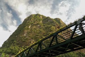 A steel bridge spans beneath a lush green mountain peak under dramatic cloudy skies in a tropical landscape.