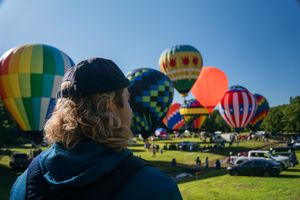 Person with long hair watching colorful hot air balloons launch at a sunny outdoor festival.