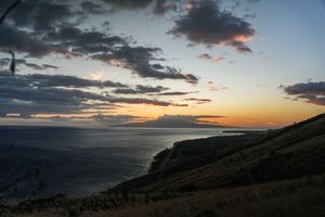 Dramatic coastal sunset with vibrant orange horizon, dark clouds, and ocean views from a clifftop hillside.