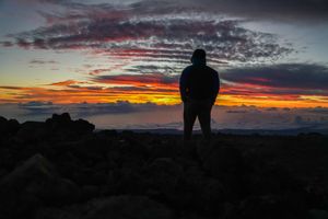 Silhouette of a person standing on rocky terrain against a vibrant sunset sky with colorful clouds over the ocean.
