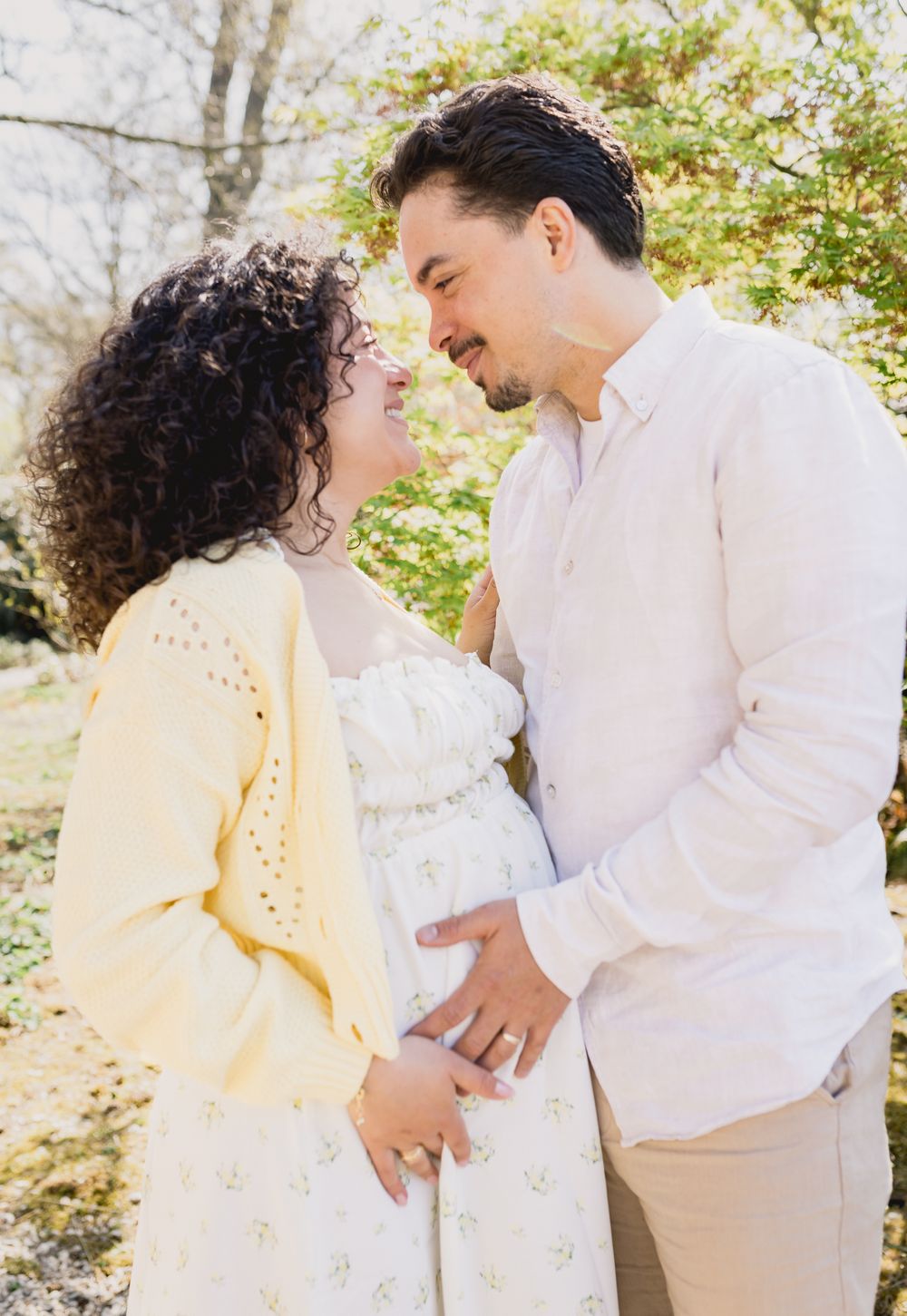 Expecting couple shares tender moment outdoors, woman in yellow cardigan and floral dress, man in white linen shirt, cradling baby bump.
