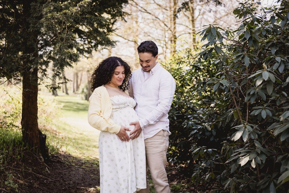 Couple sharing a tender moment outdoors, woman in floral dress cradling baby bump, man in white shirt, surrounded by lush greenery in Lisse.