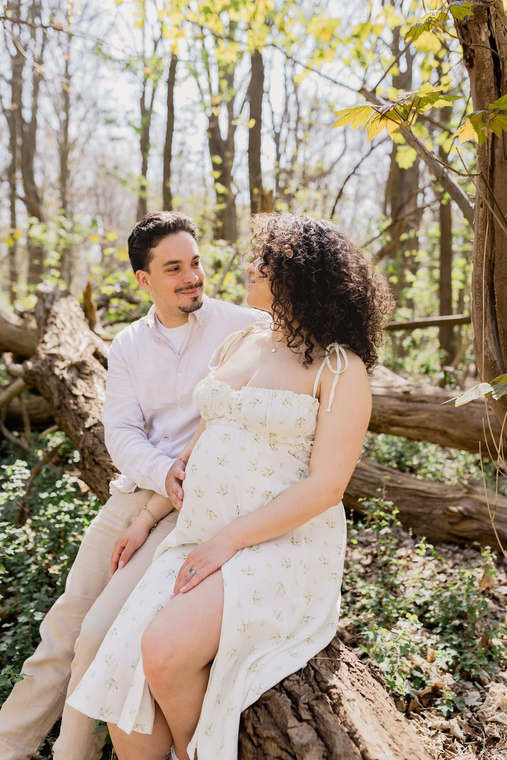 Candid portrait of pregnant couple in summer attire sitting among trees looking at each other