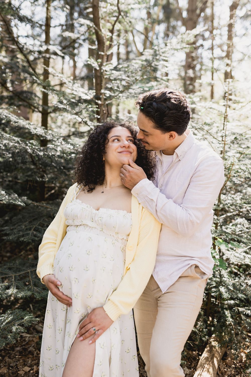 Couple in forest, pregnant woman in floral dress, man tenderly touching her face, maternity photo shoot.