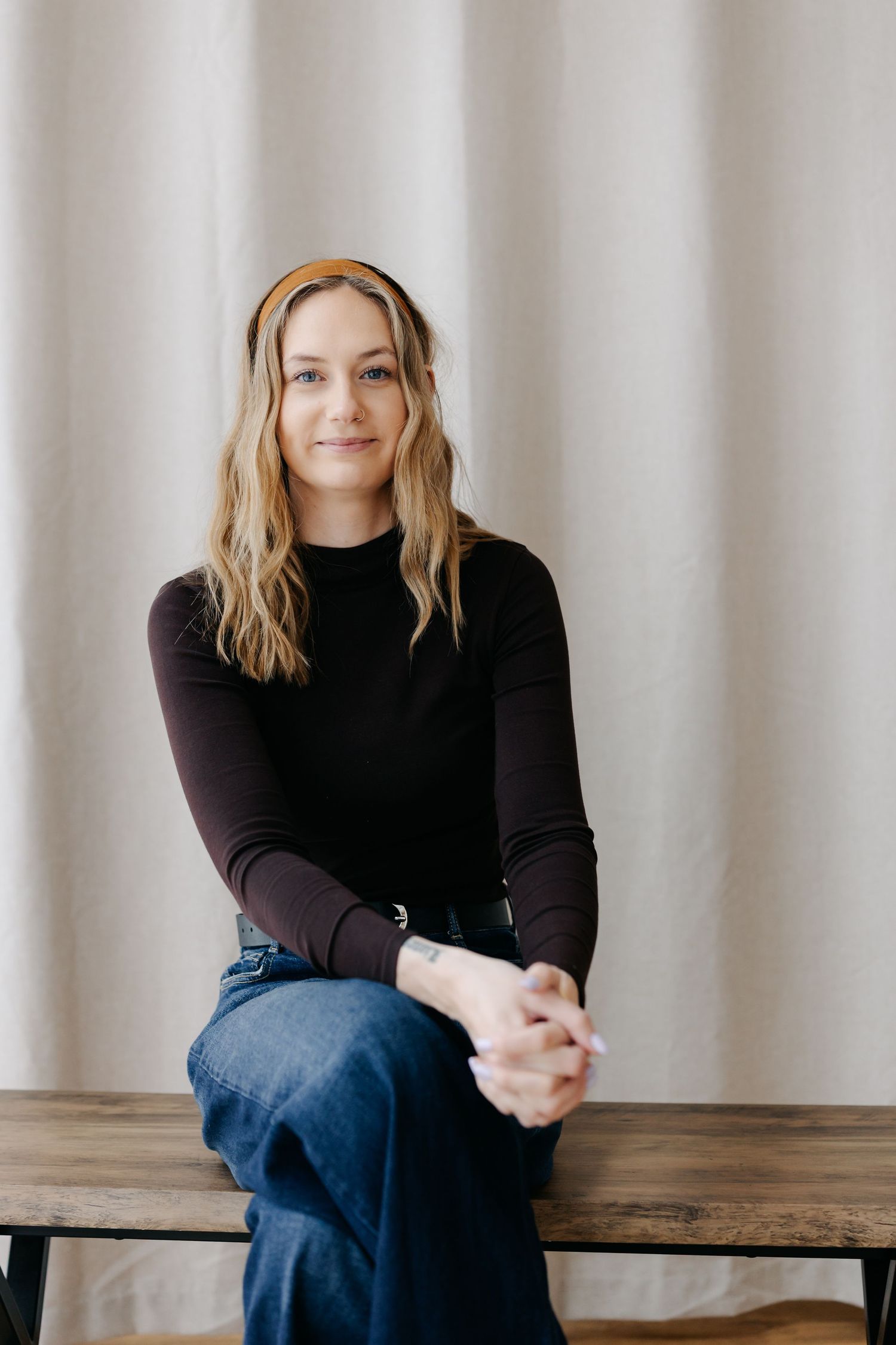 A young woman with blonde hair and a headband sits on a wooden bench, wearing a black top and jeans.