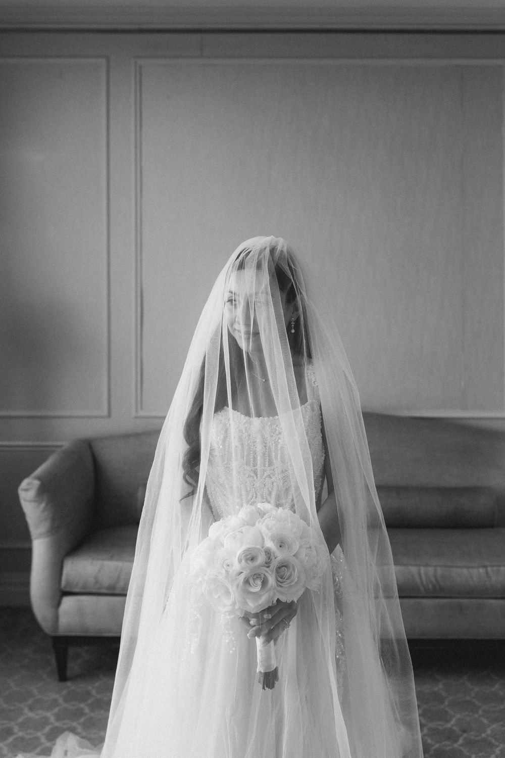 Black and white photo of a bride in a lace gown, veil over face, holding a rose bouquet, standing before a sofa.