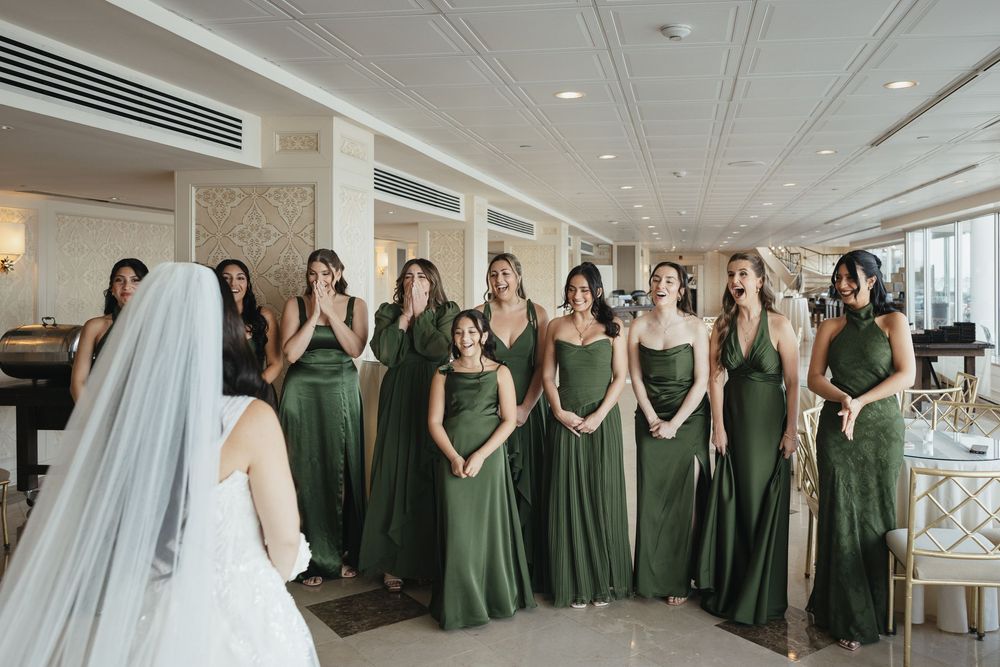 Bridesmaids in green dresses react with awe as bride reveals her wedding gown during a first look moment.