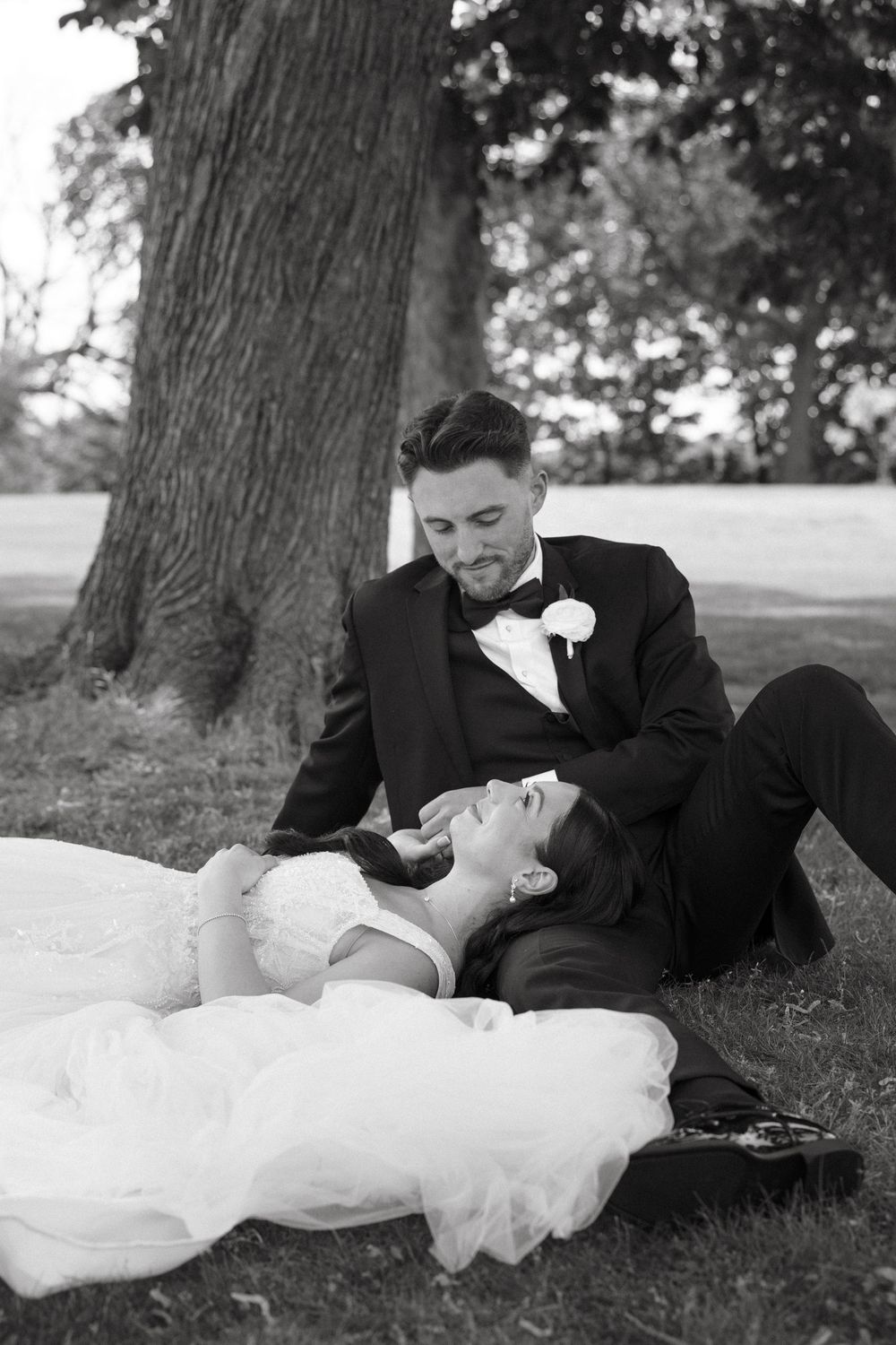 Black and white photo of a bride and groom sitting under a tree, with the bride lying in the groom's lap.