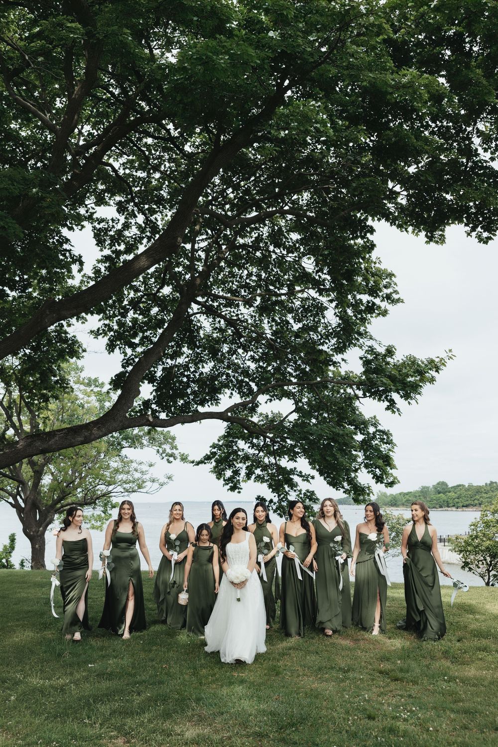 Bride in white gown poses with bridesmaids in olive green dresses under a large tree near a lake.