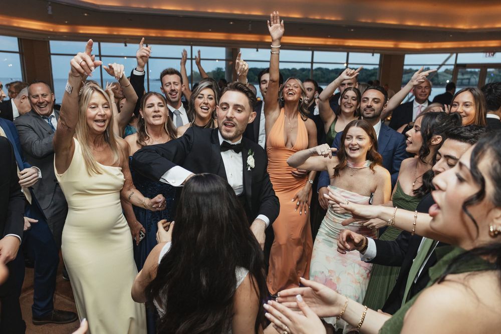 Groom in tuxedo celebrates with joyful wedding guests dancing together at elegant indoor wedding reception.