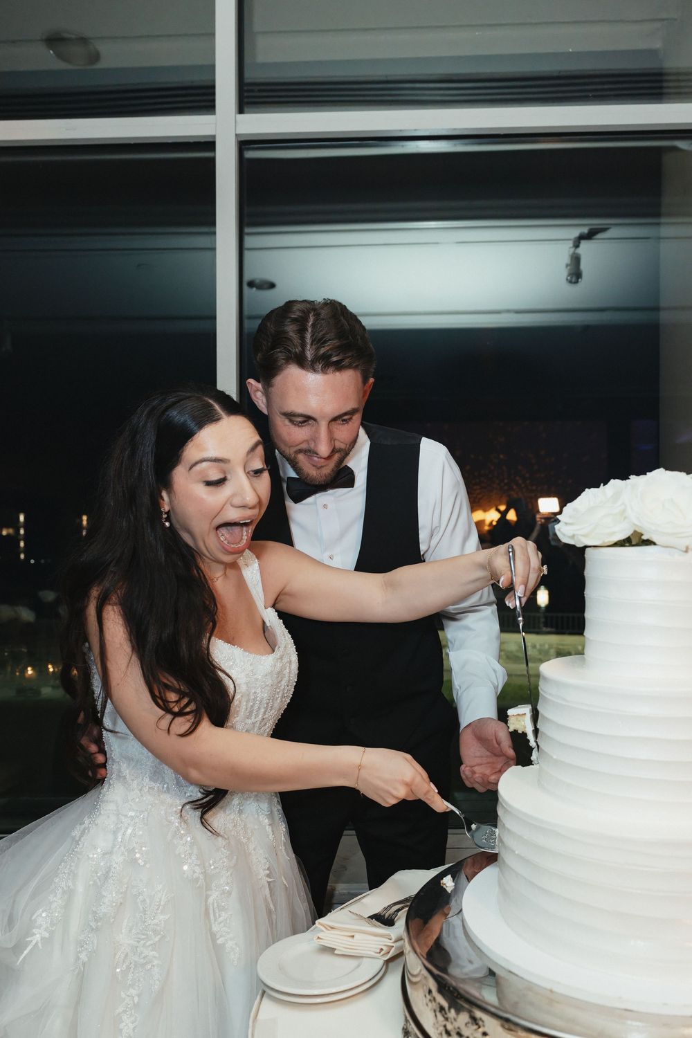 Bride and groom joyfully cutting their white tiered wedding cake together at their elegant evening reception.