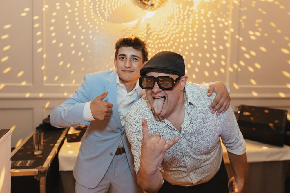 Two men posing at a party, one in a light suit giving thumbs up, other in sunglasses making rock sign with tongue out.