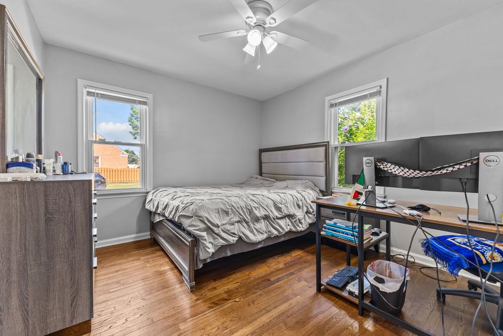 Bright bedroom with hardwood floors, ceiling fan, two windows, and a desk with dual monitors.
