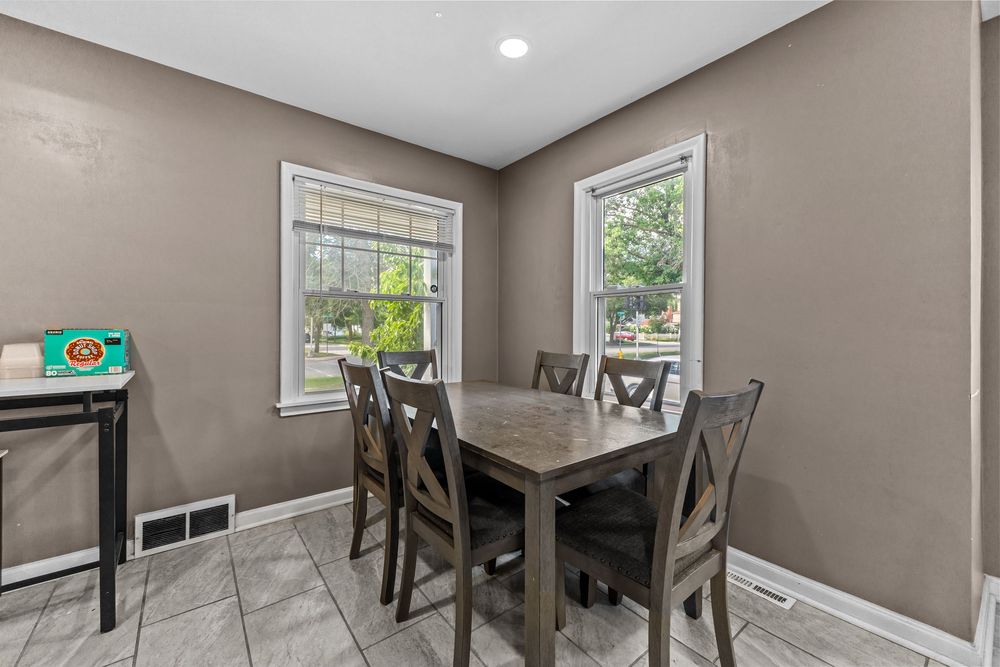 Dining area with tile floors, wooden table and chairs, two windows, and neutral taupe walls.