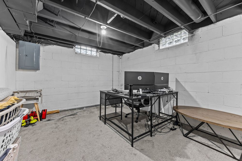 Unfinished basement with concrete floors, exposed ceiling, glass block windows, and a desk setup.