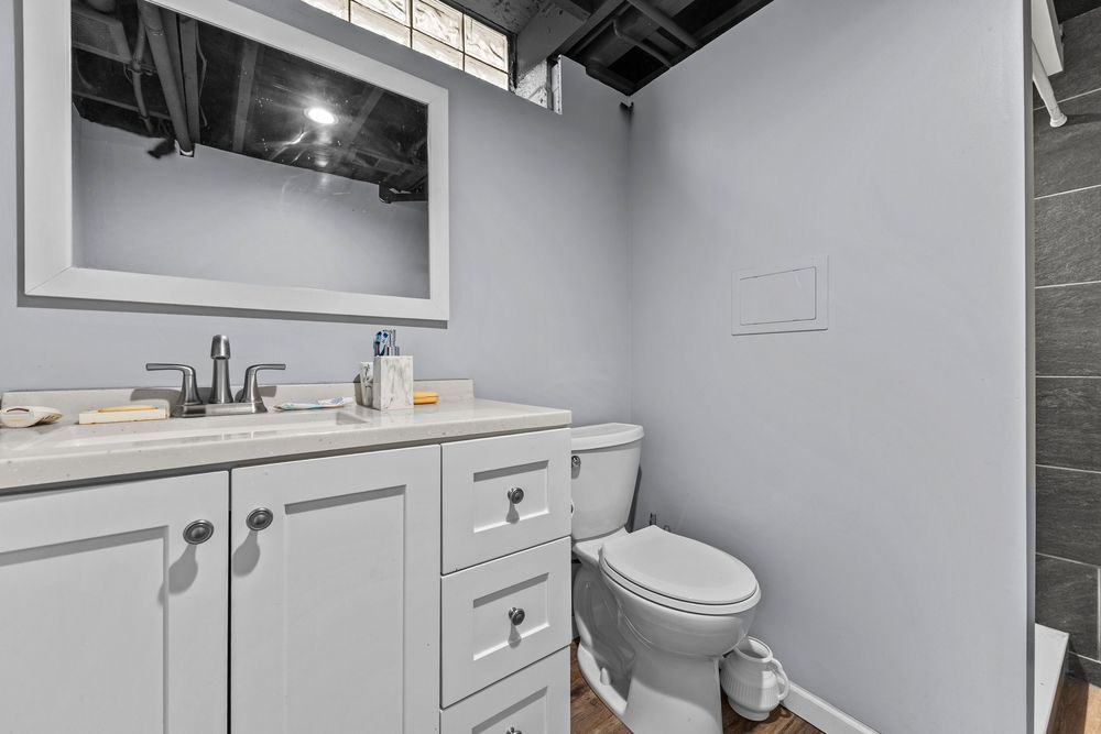 Basement half bathroom with gray vanity, large mirror, toilet, and glass block window accents.