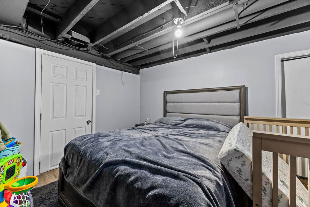 Basement bedroom with gray bedding, upholstered headboard, exposed black ceiling, white door, and a crib in the corner.