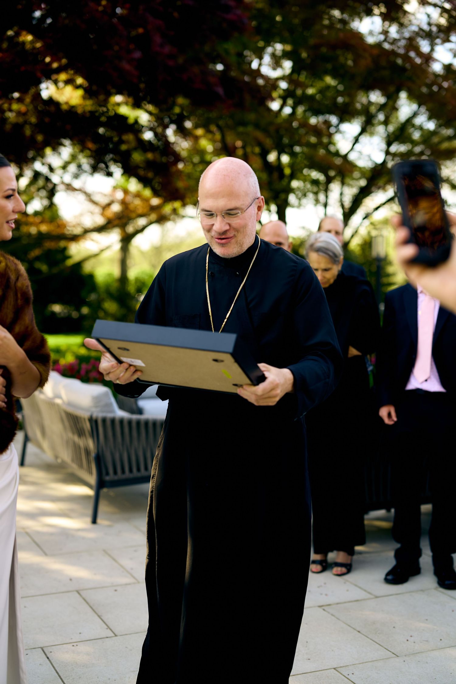 Bald priest in black robes smiling while holding a framed gift outdoors at a garden ceremony.