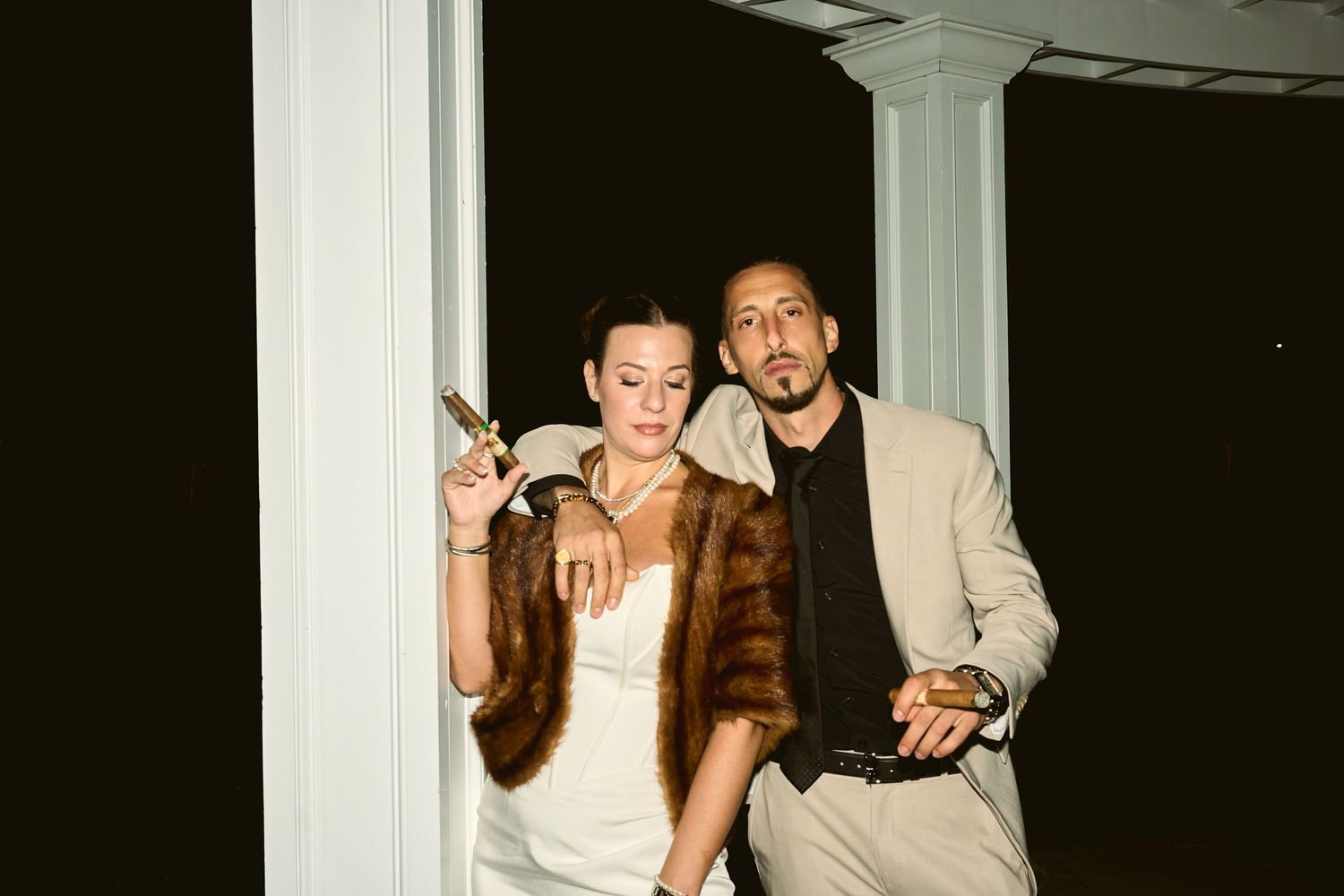 Elegant couple posing at night, woman in white dress with fur stole holding cigarette, man in beige suit.