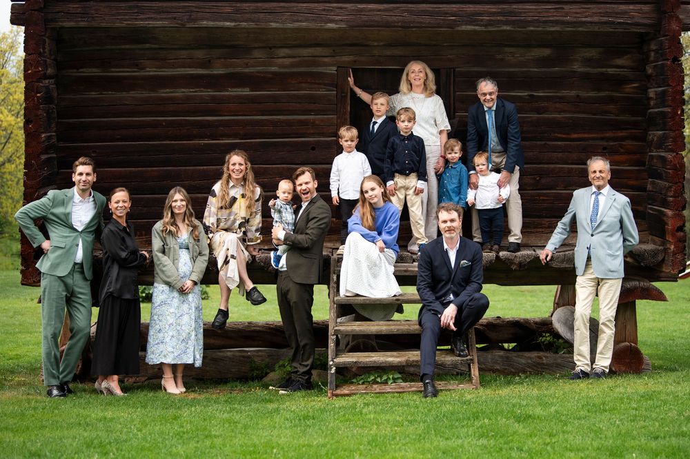 Large family group portrait outdoors in front of a rustic wooden cabin, dressed in elegant formal attire.