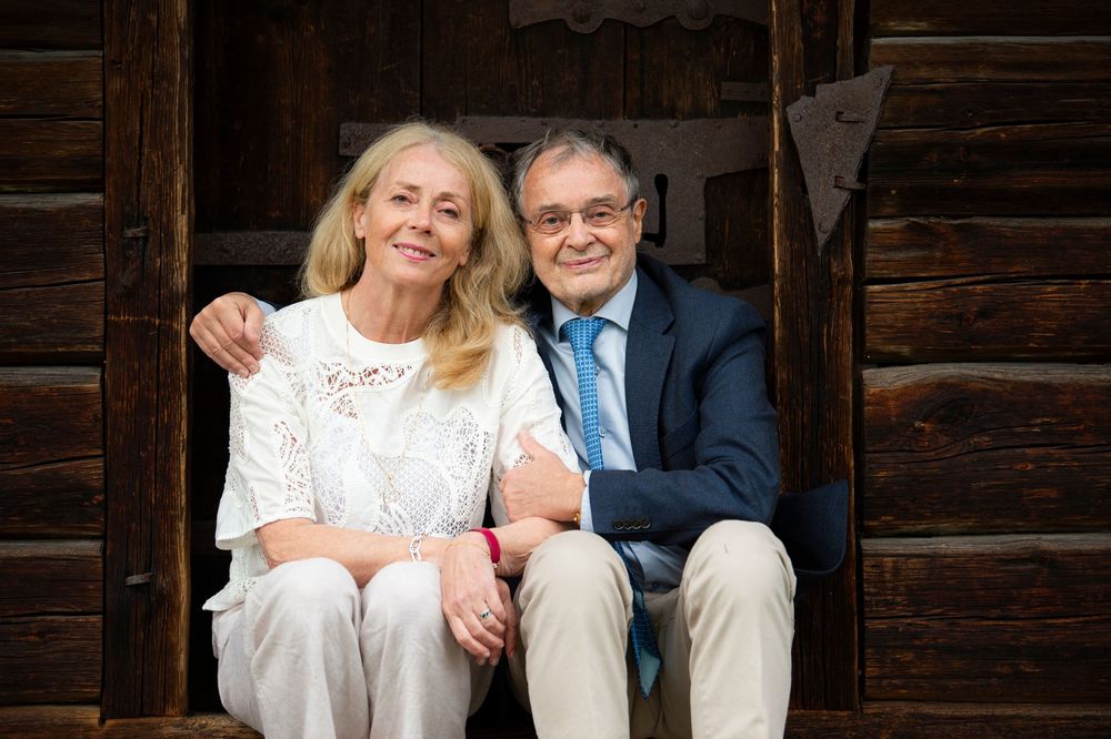 Smiling elderly couple sitting together in front of a rustic wooden cabin wall.
