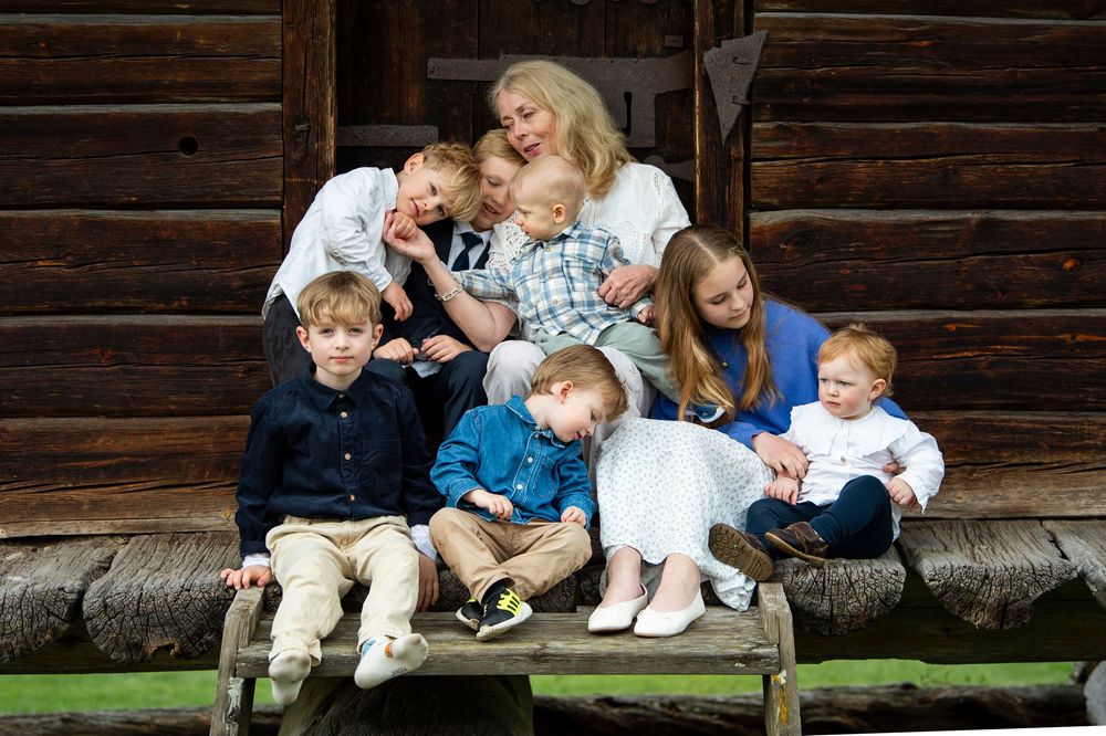Grandmother surrounded by seven grandchildren in a candid, joyful group photo outside a log cabin.