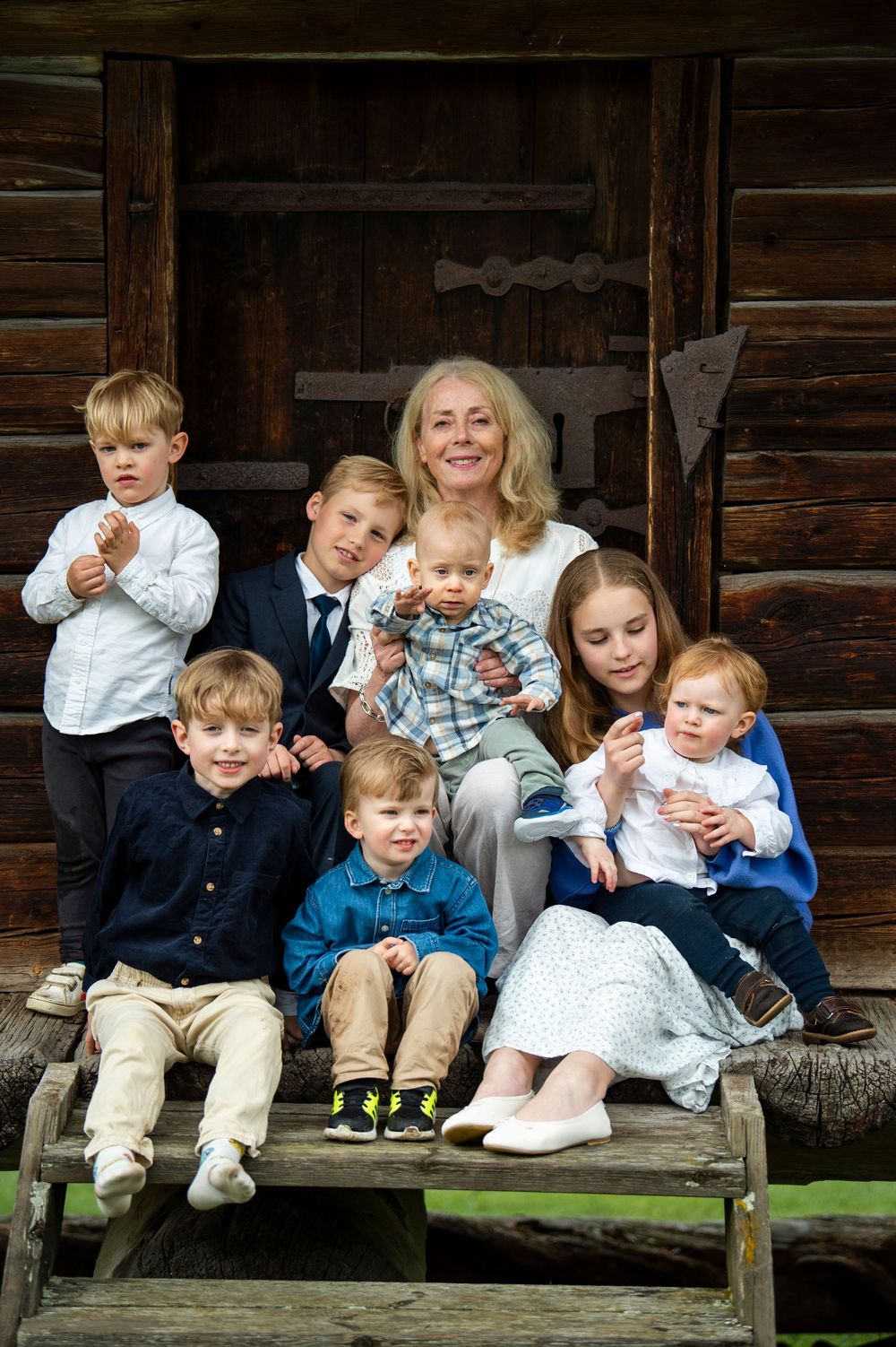 A grandmother poses with seven young grandchildren on rustic wooden steps in front of a log cabin.