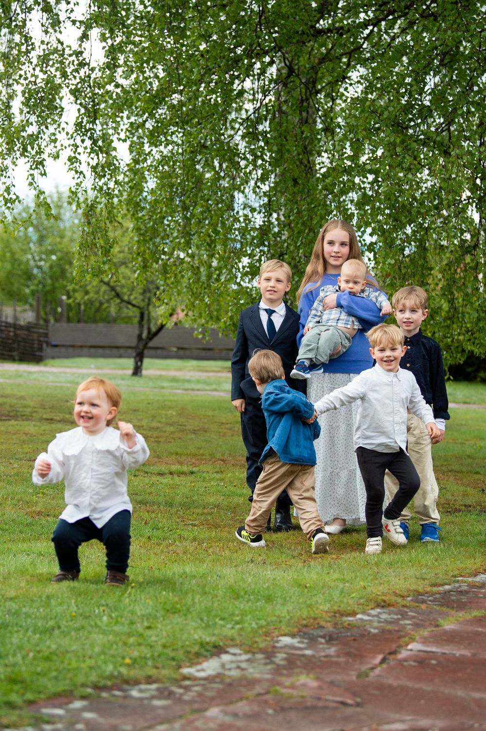 A teenage girl holds a baby while several young boys play and run around her in a lush green park.