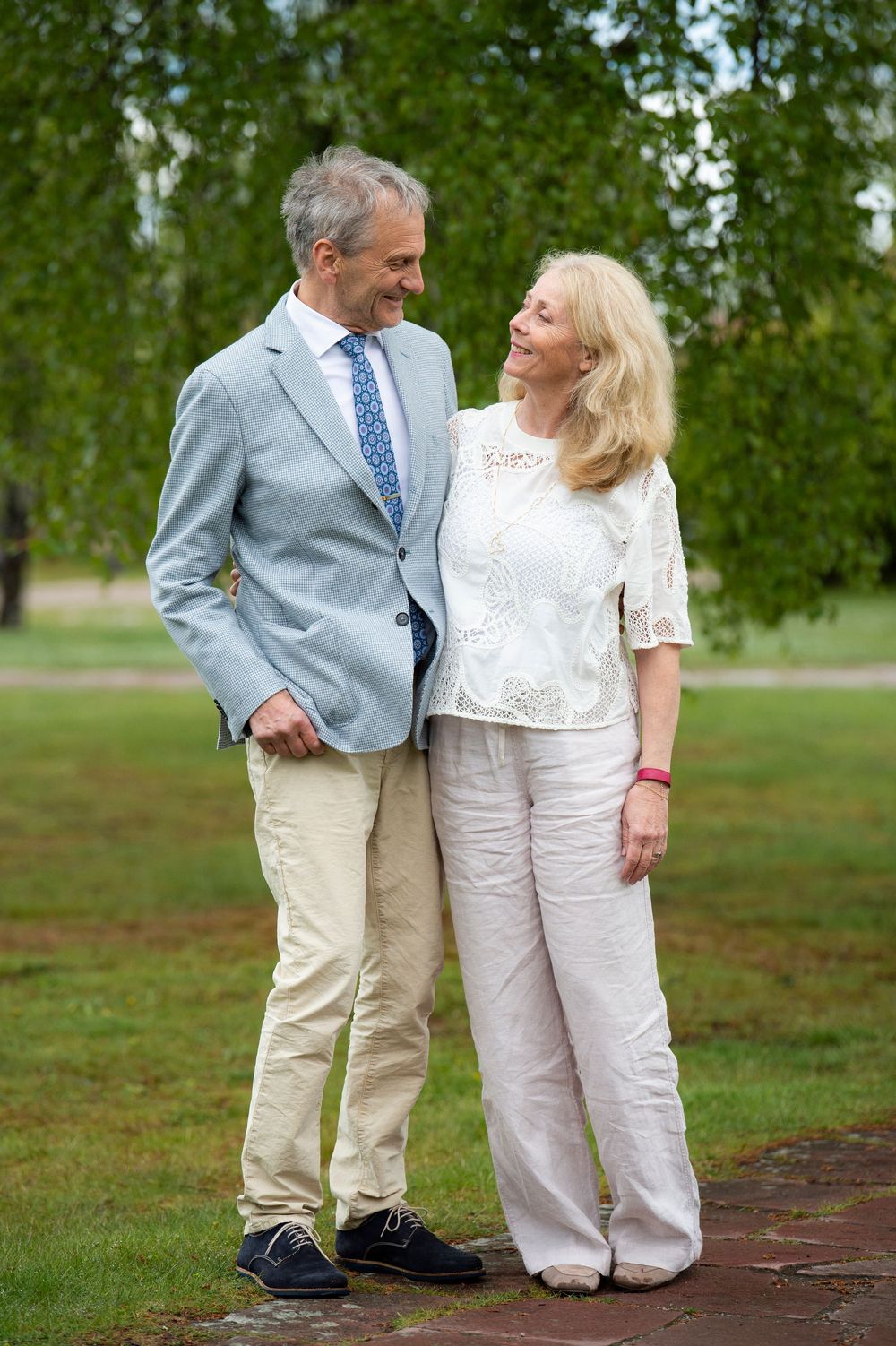 A smiling older couple in elegant attire share a loving glance while standing in a green park.