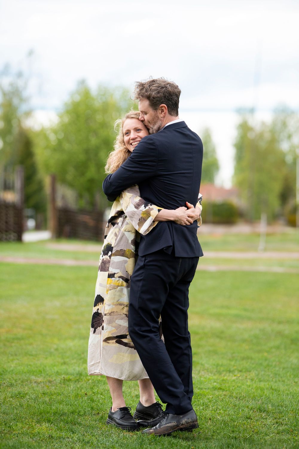 A man in a black suit and a woman in a patterned dress share a warm embrace on a green lawn.