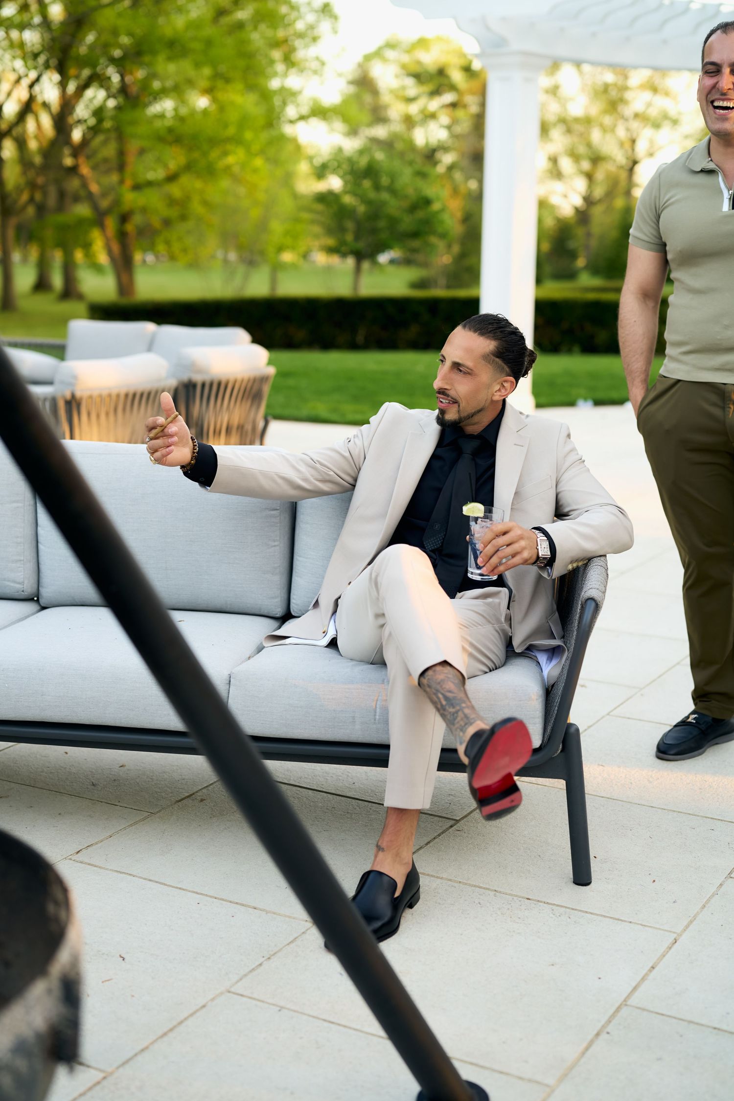 A stylish man in a beige suit sits on an outdoor sofa, holding a drink and gesturing, with lush greenery in the background.