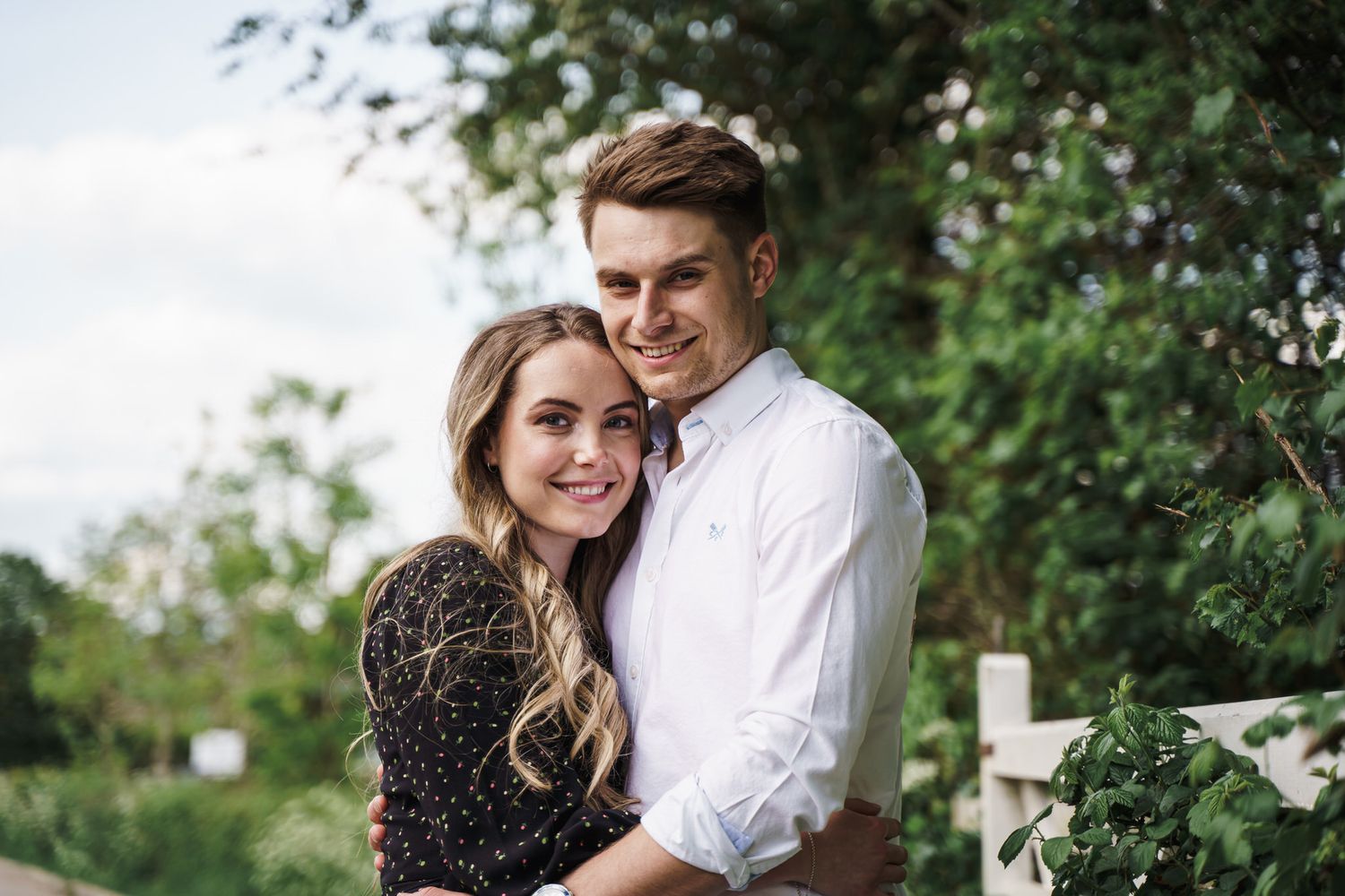 A smiling young couple embraces outdoors, surrounded by lush green trees and a white fence.