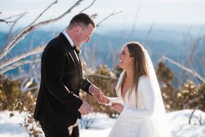 Bride and groom holding hands and smiling during winter wedding ceremony on snowy mountain summit.