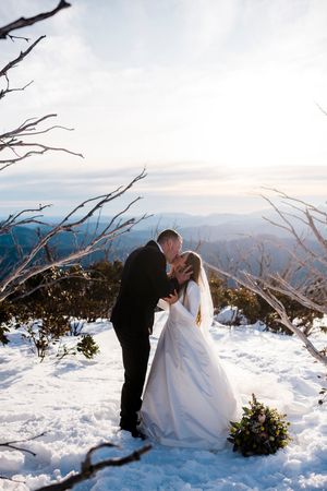 Bride and groom share a kiss on a snowy mountain summit, surrounded by bare trees and panoramic views.