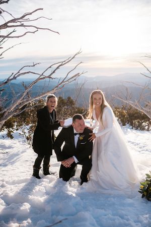 Bride and groom laughing in snow on mountain top with wedding guest, snowy winter wedding celebration outdoors.