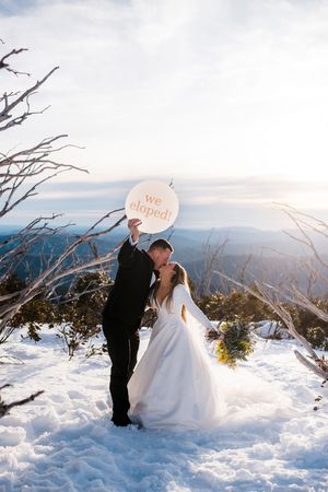 Eloping couple kiss on snowy mountain peak holding a round "we eloped" sign with scenic backdrop.