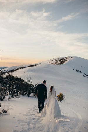 Bride and groom walking hand in hand through snowy mountain landscape at sunset, bride holding yellow bouquet with long veil flowing.