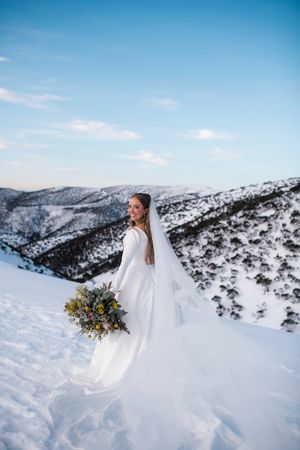 Bride in white gown and veil holds colorful bouquet on snowy mountain slope with scenic winter landscape backdrop.