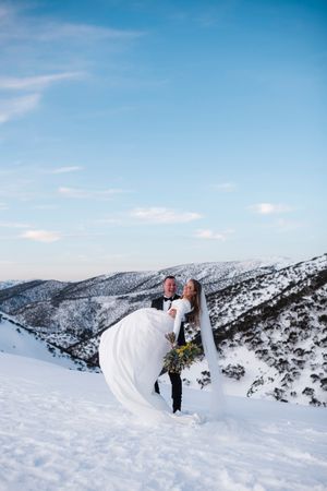 Bride and groom embrace on snowy mountain peak in winter wedding attire, with snow-covered hills and blue sky behind them.