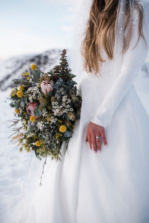 Bride in white gown holding winter bouquet with protea, eucalyptus, and yellow flowers against snowy mountain backdrop.